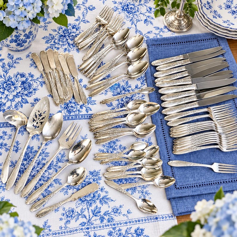 May include: A large collection of silver-toned flatware featuring ornate floral detailing on the handles. The set includes various sizes of forks, knives, spoons, and serving utensils arranged on a blue and white floral patterned tablecloth.
