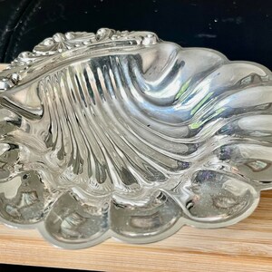 May include: A silver, shell-shaped dish with a detailed, ornate design. The dish is made of polished metal and has a reflective surface.