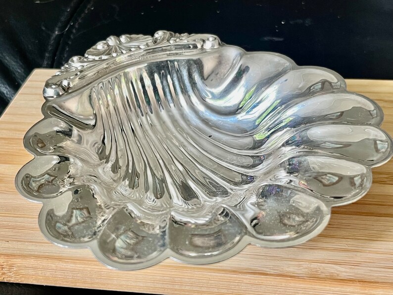 May include: A silver, shell-shaped dish with a detailed, ornate design. The dish is made of polished metal and has a reflective surface.