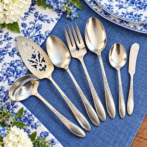 May include: A set of six silver-toned metal utensils including a ladle, a pierced cake server, a large spoon, a three-tined fork, a small spoon, and a butter knife, all featuring a matching embossed floral pattern on the handles.