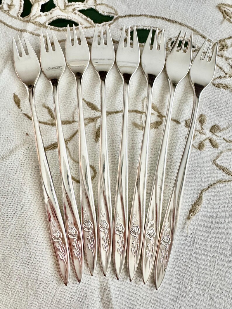 Op de afbeelding: Een verzameling van acht zilveren vorken. Elke vork heeft een ontwerp met drie tanden en een lange, slanke steel. De stelen zijn versierd met een rozen- en bladerenpatroon. De vorken zijn gerangschikt op een wit oppervlak met een bloemenmotief.