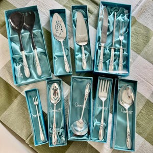 May include: A collection of silver-colored serving utensils, including spoons, forks, and knives, displayed in their original blue boxes. The utensils have ornate handles and are arranged on a green and white checkered surface.
