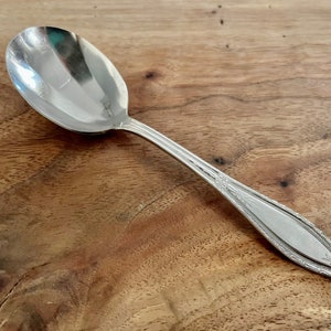 May include: A silver spoon with an ornate design on the handle. The spoon is on a wooden surface.