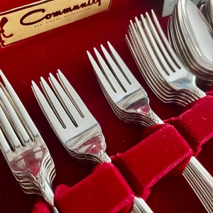 May include: A set of silver silverware, including forks and spoons, arranged in a red velvet-lined box. The forks and spoons are neatly organized, with a decorative pattern on the handles. The box has a gold label that reads "Community The Finest Silverplate."
