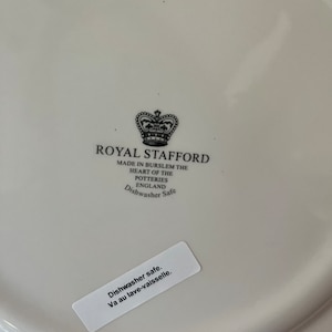 Op de afbeelding: Een witte keramische bordbodem met een zwart gedrukt Royal Stafford kroonlogo en de tekst Made in Burslem The Heart of the Potteries England Dishwasher Safe. Een witte rechthoekige sticker bevat de tekst Dishwasher safe. Va au lave-vaisselle.