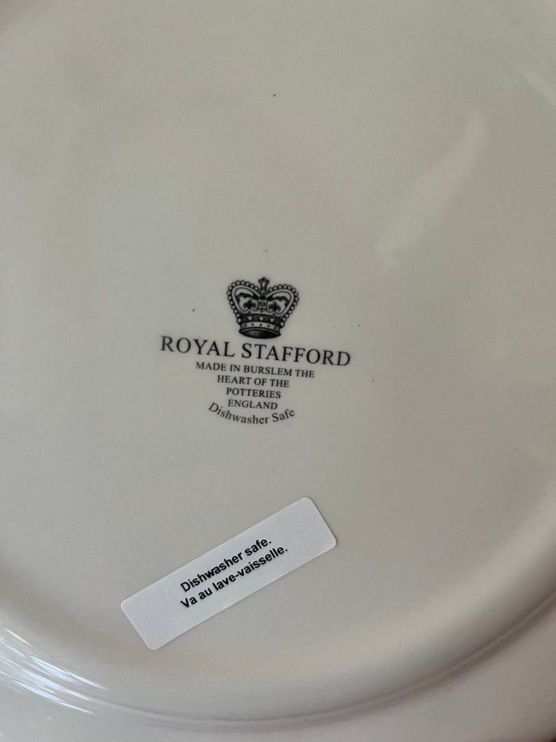 Op de afbeelding: Een witte keramische bordbodem met een zwart gedrukt Royal Stafford kroonlogo en de tekst Made in Burslem The Heart of the Potteries England Dishwasher Safe. Een witte rechthoekige sticker bevat de tekst Dishwasher safe. Va au lave-vaisselle.
