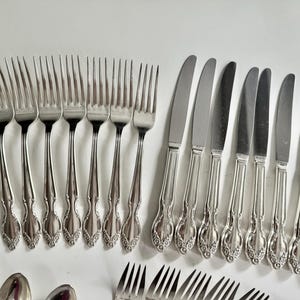 May include: A collection of silver-colored forks and knives. The forks have four tines and ornate handles. The knives have a smooth blade and a matching handle design. The silverware is arranged on a white surface.