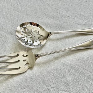 May include: Silver serving spoon with a pierced floral design and a matching serving fork. Both utensils have long, slender handles with a decorative element at the end. The spoon has a rounded bowl, and the fork has three tines.