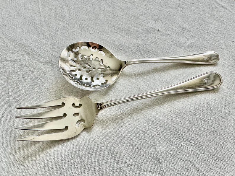 May include: Silver serving spoon with a pierced floral design and a matching serving fork. Both utensils have long, slender handles with a decorative element at the end. The spoon has a rounded bowl, and the fork has three tines.