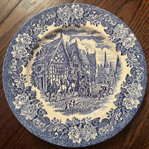 May include: A round ceramic plate featuring a blue and white transferware pattern. The center depicts a historical street scene with a horse-drawn carriage, timber-framed buildings, and a dog. The rim is decorated with a detailed floral border.