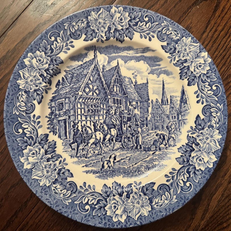 May include: A round ceramic plate featuring a blue and white transferware pattern. The center depicts a historical street scene with a horse-drawn carriage, timber-framed buildings, and a dog. The rim is decorated with a detailed floral border.