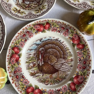 Peut inclure: Une assiette en céramique blanche avec une bordure brune et verte représentant une dinde, des pommes et des fleurs. Le centre de l'assiette présente une illustration détaillée d'une dinde dans un cadre rural.