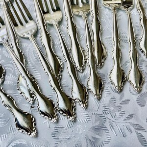 May include: A collection of silver forks with ornate, curved handles. The forks are arranged diagonally across a white, patterned tablecloth. The tines of the forks are visible, and the silverware has a shiny, reflective surface.