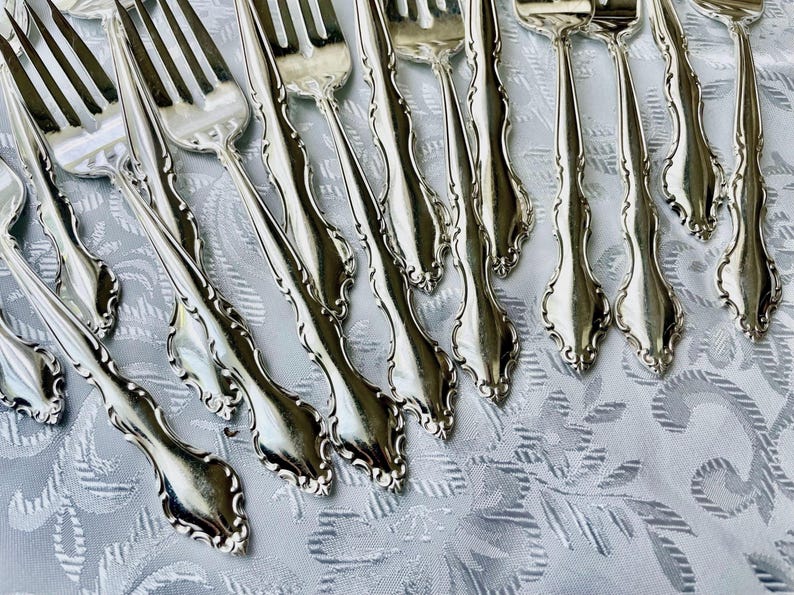 May include: A collection of silver forks with ornate, curved handles. The forks are arranged diagonally across a white, patterned tablecloth. The tines of the forks are visible, and the silverware has a shiny, reflective surface.