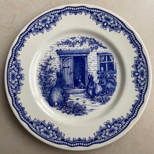 Puede incluir: Un plato redondo de cer&aacute;mica con un patr&oacute;n de transferencia azul y blanco. El centro representa una casa de ladrillo con varios conejos en primer plano. El borde est&aacute; decorado con un dise&ntilde;o repetitivo de flores y volutas en azul oscuro.