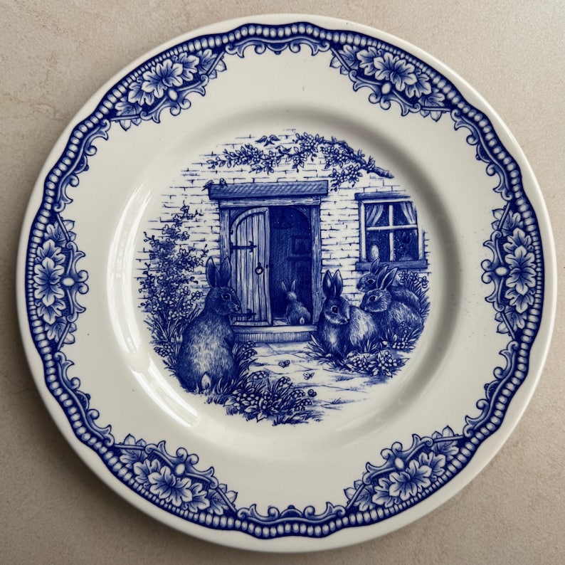 Puede incluir: Un plato redondo de cer&aacute;mica con un patr&oacute;n de transferencia azul y blanco. El centro representa una casa de ladrillo con varios conejos en primer plano. El borde est&aacute; decorado con un dise&ntilde;o repetitivo de flores y volutas en azul oscuro.