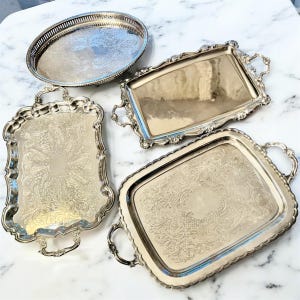 Pu&ograve; includere: Una collezione di quattro vassoi da portata color argento. Uno &egrave; ovale con un bordo traforato, mentre gli altri tre sono rettangolari con motivi floreali in rilievo, bordi smerlati e manici decorativi su entrambi i lati.