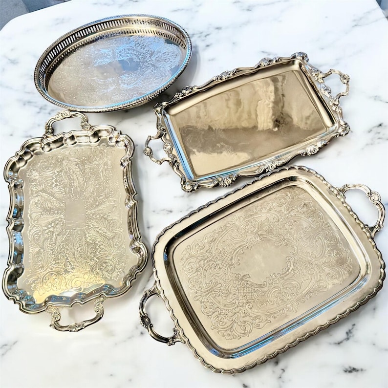 Pu&ograve; includere: Una collezione di quattro vassoi da portata color argento. Uno &egrave; ovale con un bordo traforato, mentre gli altri tre sono rettangolari con motivi floreali in rilievo, bordi smerlati e manici decorativi su entrambi i lati.