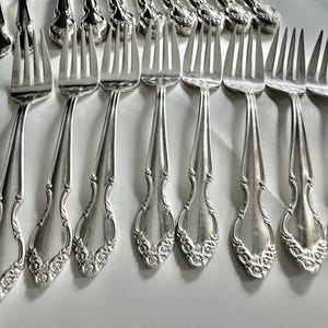 May include: A collection of antique silver forks with ornate floral detailing on the handles. The forks are arranged in rows, showcasing their elegant design and polished finish. The tines are evenly spaced.