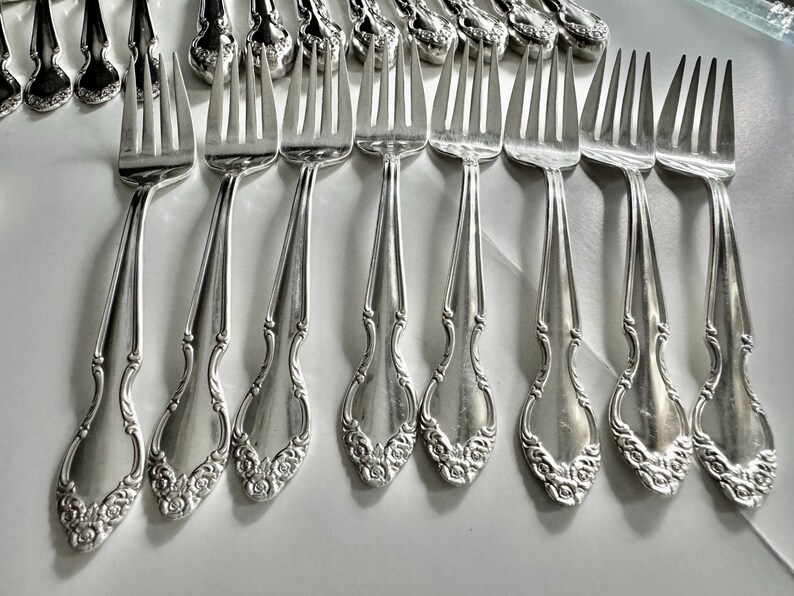 May include: A collection of antique silver forks with ornate floral detailing on the handles. The forks are arranged in rows, showcasing their elegant design and polished finish. The tines are evenly spaced.