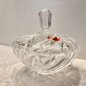 May include: A clear, cut-glass candy dish with a matching lid featuring a faceted finial. The bowl has a radial, starburst-like pattern etched into the glass. A small, red and gold sticker is adhered to the top surface of the glass lid.