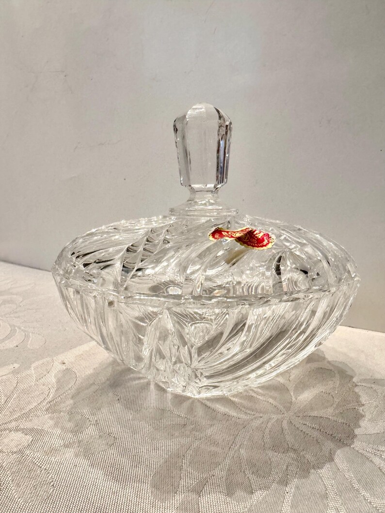 May include: A clear, cut-glass candy dish with a matching lid featuring a faceted finial. The bowl has a radial, starburst-like pattern etched into the glass. A small, red and gold sticker is adhered to the top surface of the glass lid.