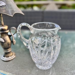 Puede incluir: Una jarra de cristal transparente con asa y boquilla. La jarra tiene un dise&ntilde;o de cristal tallado. Una l&aacute;mpara plateada est&aacute; en el fondo. La jarra est&aacute; sobre una superficie verde claro.