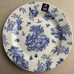 Puede incluir: Un plato redondo de cer&aacute;mica con base color crema y un patr&oacute;n toile azul que presenta escenas pastorales, &aacute;rboles, p&aacute;jaros y mariposas. Una peque&ntilde;a pegatina rectangular en la parte superior muestra el texto Royal Wares Est 1792.