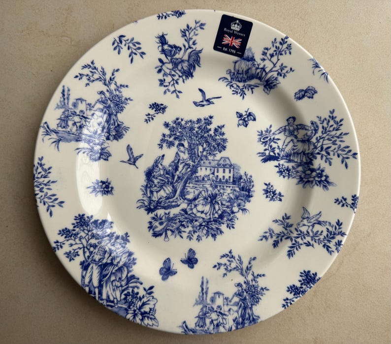 Puede incluir: Un plato redondo de cer&aacute;mica con base color crema y un patr&oacute;n toile azul que presenta escenas pastorales, &aacute;rboles, p&aacute;jaros y mariposas. Una peque&ntilde;a pegatina rectangular en la parte superior muestra el texto Royal Wares Est 1792.