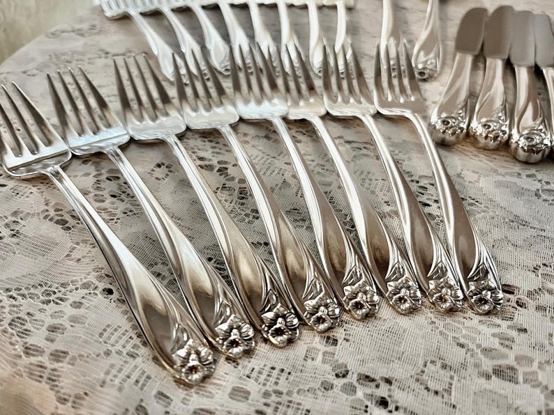May include: A set of silver forks with ornate floral designs on the handles. The forks are arranged in a row on a white lace tablecloth.