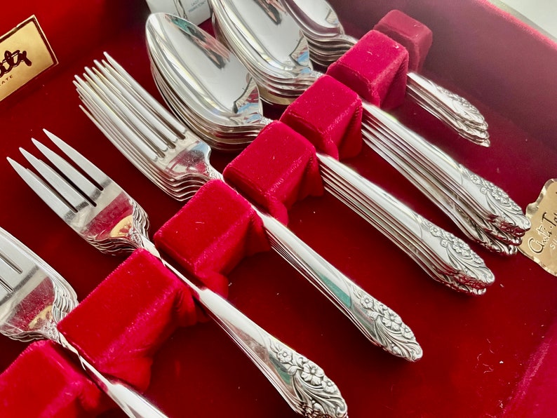 May include: A set of silver-plated flatware, including forks and spoons, arranged in a red velvet-lined box. The silverware features a floral design on the handles. The set is ideal for formal dining occasions.