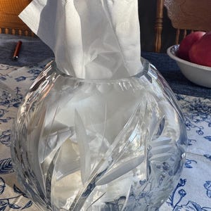 May include: A clear, spherical glass tissue holder featuring intricate, etched leaf and floral patterns across its surface. The container is filled with white paper tissues that extend from the top opening, showcasing a polished, reflective finish.