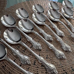 May include: A collection of silver serving spoons with ornate, floral-inspired handle designs. The spoons are arranged on a textured, brown surface, reflecting light. The spoons are likely part of a formal dining set.