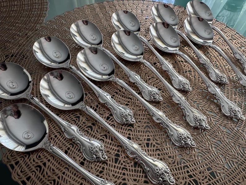 May include: A collection of silver serving spoons with ornate, floral-inspired handle designs. The spoons are arranged on a textured, brown surface, reflecting light. The spoons are likely part of a formal dining set.