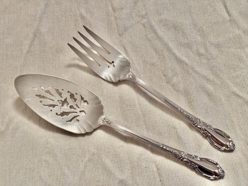 K&ouml;nnte beinhalten: Ein silberner Kuchenspachtel und eine Gabel mit verzierten Griffen und dekorativen Ausschnitten. Der Spachtel hat ein Blattdesign und die Gabel ein Blumendesign.