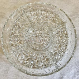 May include: Clear glass serving dish with a starburst pattern. The dish is divided into five sections, with a central circular area and four pie-shaped compartments. The rim has a decorative edge. Ideal for serving snacks or appetizers.