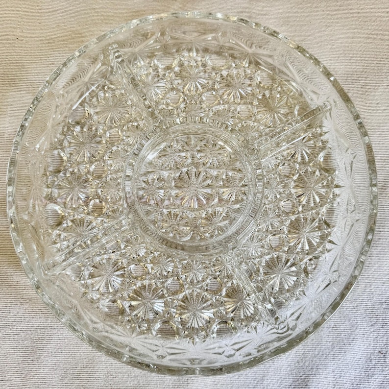 May include: Clear glass serving dish with a starburst pattern. The dish is divided into five sections, with a central circular area and four pie-shaped compartments. The rim has a decorative edge. Ideal for serving snacks or appetizers.