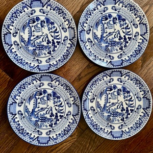 K&ouml;nnte beinhalten: Vier passende blau-wei&szlig;e Porzellanteller mit einem detaillierten, traditionellen Design. Jeder Teller zeigt eine zentrale Szene, umgeben von komplizierten Mustern und einem blauen Rand. Die Teller sind auf einer Holzoberfl&auml;che angeordnet.