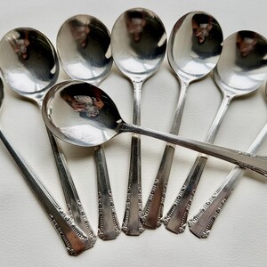 May include: Eight silver soup spoons with ornate handles. The spoons are arranged in a fan shape, with the handles pointing outwards.