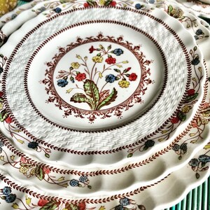 Op de afbeelding: Een set van drie witte keramische borden met een geweven patroon en een bloemenontwerp in tinten rood, groen, blauw en geel. De borden hebben een gegolfde rand.