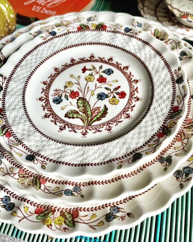 Op de afbeelding: Een set van drie witte keramische borden met een geweven patroon en een bloemenontwerp in tinten rood, groen, blauw en geel. De borden hebben een gegolfde rand.