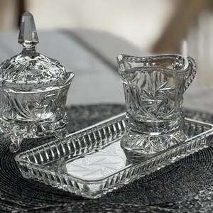 May include: A clear pressed glass set consisting of a lidded sugar bowl with three feet, a creamer pitcher, and a rectangular tray. Each piece features a detailed starburst and geometric pattern with scalloped edges and a faceted finial on the lid.