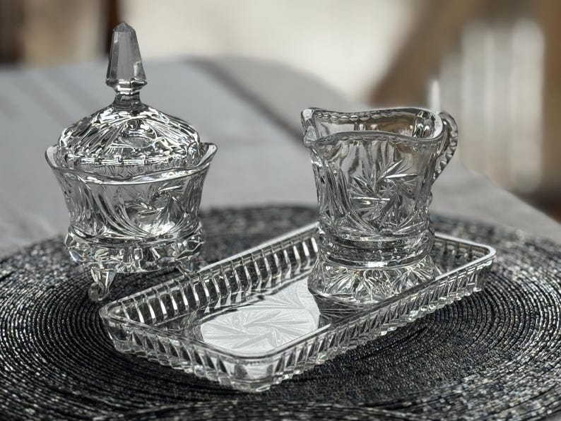 May include: A clear pressed glass set consisting of a lidded sugar bowl with three feet, a creamer pitcher, and a rectangular tray. Each piece features a detailed starburst and geometric pattern with scalloped edges and a faceted finial on the lid.