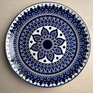 May include: A round ceramic plate featuring a detailed, symmetrical mandala pattern in shades of deep blue and white. The design consists of concentric rings of intricate leaf-like shapes, dots, and geometric lines radiating from a solid dark center.