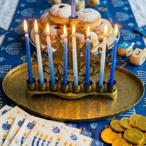 May include: A brass menorah with lit blue and white candles sits on a gold tray, surrounded by sufganiyot, dreidels, and gold coins. Napkins with menorah designs are also present, all set on a blue tablecloth.