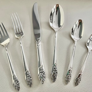 May include: A set of six silver-colored flatware pieces, including forks, a knife, and spoons. The silverware features a floral design on the handles. The pieces are arranged on a light-colored surface, reflecting the light.