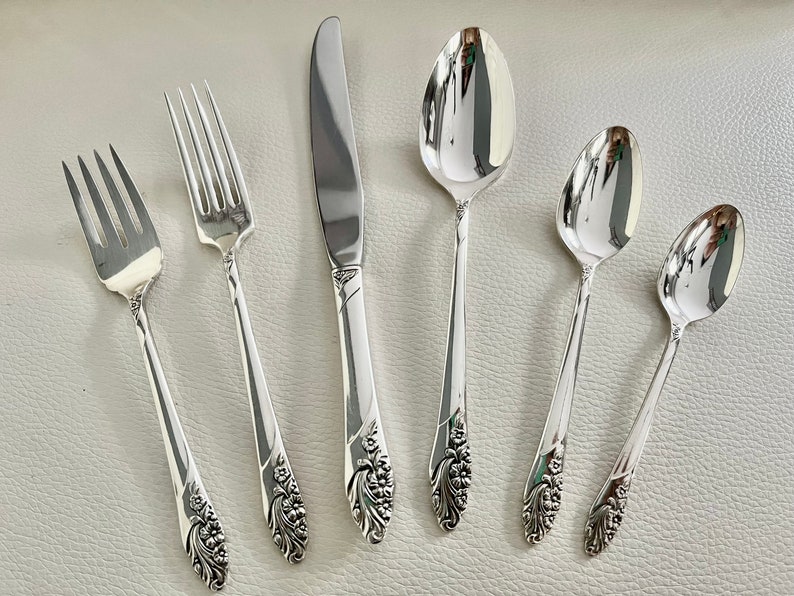 May include: A set of six silver-colored flatware pieces, including forks, a knife, and spoons. The silverware features a floral design on the handles. The pieces are arranged on a light-colored surface, reflecting the light.