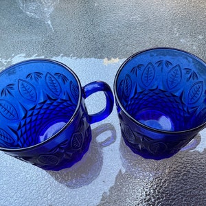 May include: Two cobalt blue glass mugs with a leaf pattern and diamond-shaped design. The mugs have handles.