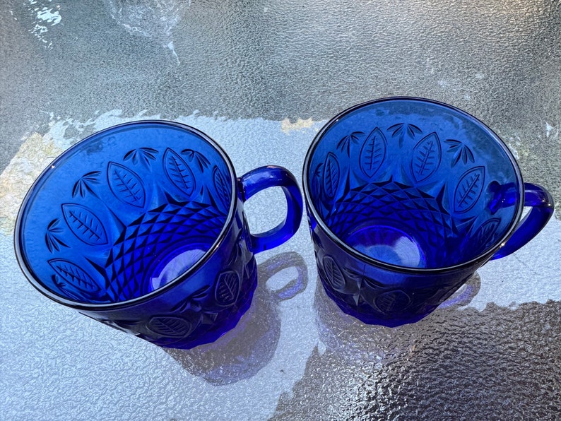 May include: Two cobalt blue glass mugs with a leaf pattern and diamond-shaped design. The mugs have handles.