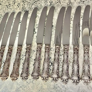 May include: A set of twelve silver-toned dinner knives with smooth, polished blades and ornate, embossed handles featuring intricate scrollwork and floral detailing. The knives are arranged in a row, showcasing the consistent design of the metal handles.